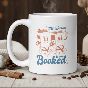 White mug with cartoon design and text 'My Weekend is all Booked' on a wooden table with coffee beans and a vase.