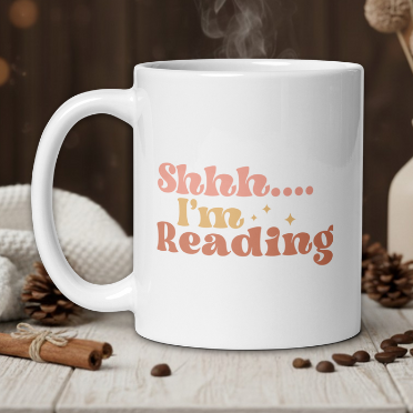 White mug with 'Shhh.... I'm Reading' text on a wooden table with coffee beans and a vase in the background.
