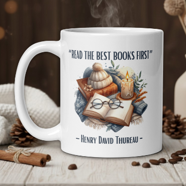White mug with book-themed design and quote on a wooden table with coffee beans and a vase.