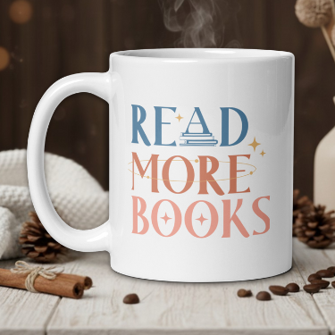 White mug with 'Read More Books' text on a wooden table with coffee beans and a vase in the background.