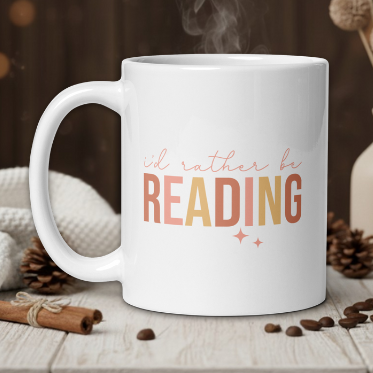 White mug with 'I'd rather be reading' text on a wooden table with coffee beans and a vase in the background.