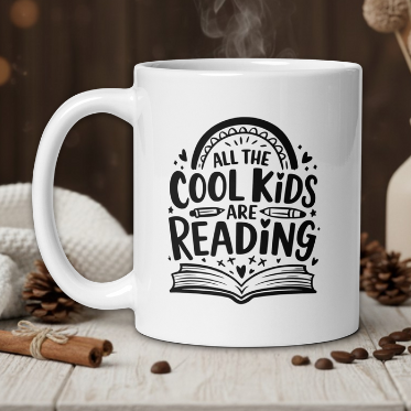 White mug with 'All the cool kids are reading' design on a wooden table with coffee beans and a vase.
