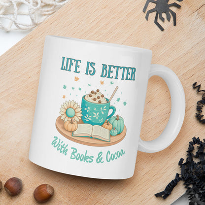 Mug with 'Life is Better with Books & Cocoa' design on a wooden board with nuts and spider decorations.