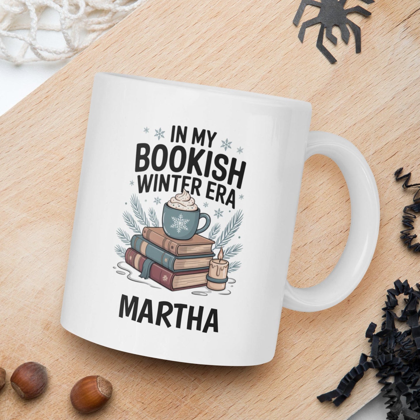 White mug with 'In my bookish winter era' design on a wooden board with nuts and spider decorations.